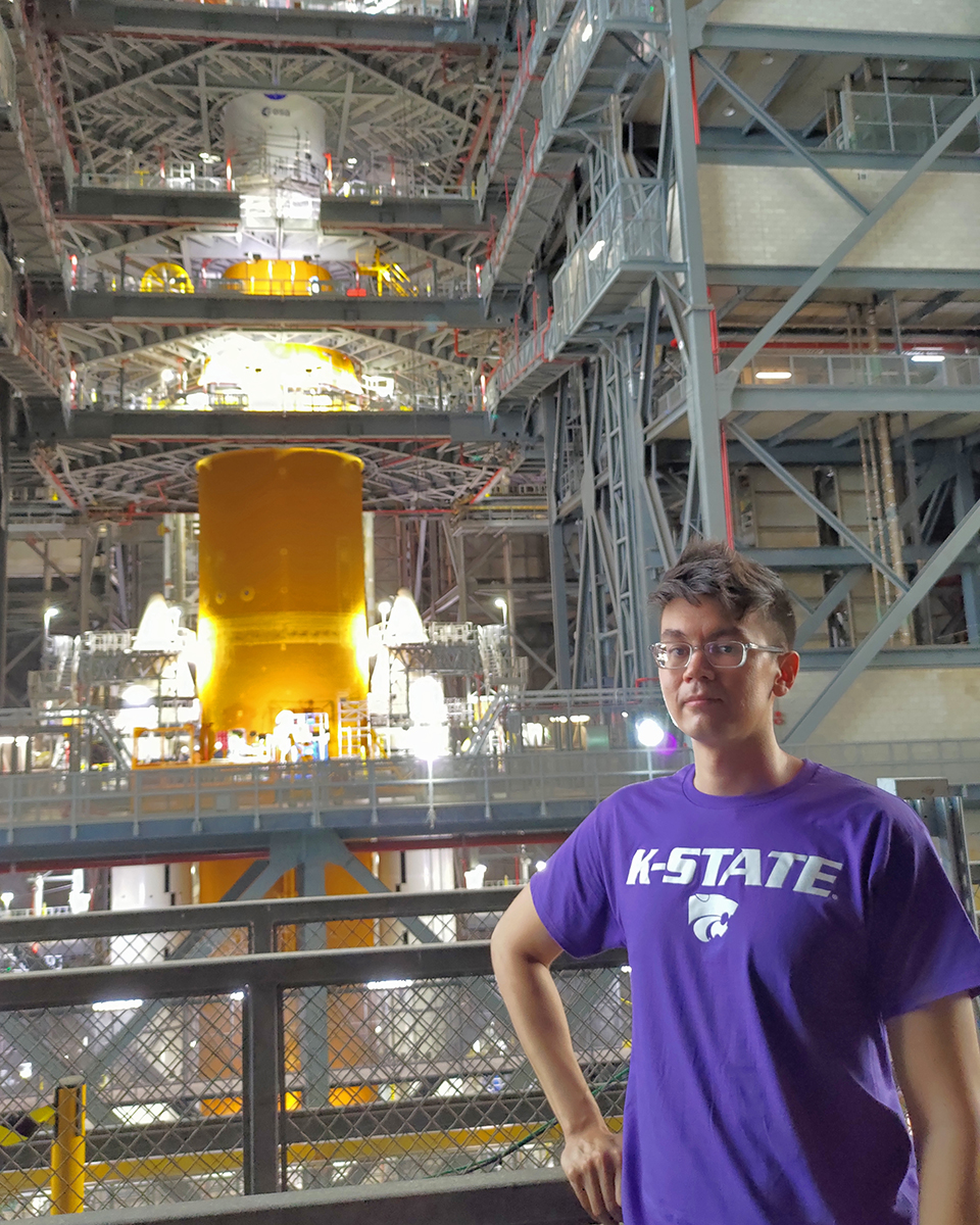 A male rocket engineer in a purple shirt with a K‑State Powercat symbol poses for a portrait in a rocket assembly tower.