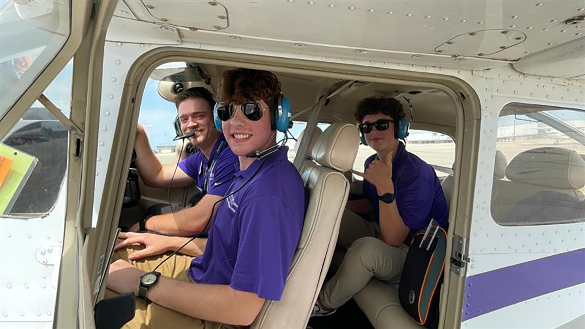 A group of high school boys sit and pose for a photo in the cockpit of a small airplane on an airport tarmac.