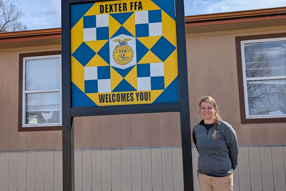 A woman in a grey long-sleeved shirt stands in front of a yellow and blue Dexter FFA Welcomes You sign