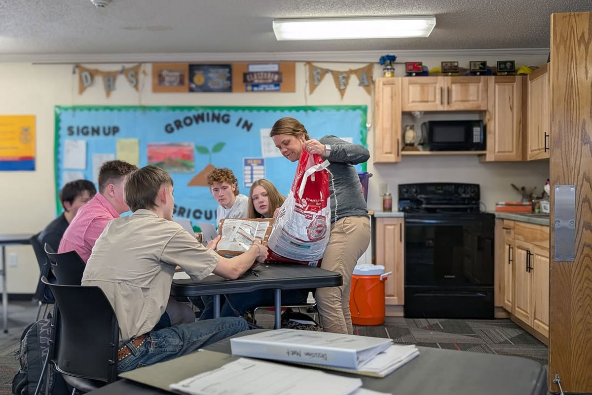 A teacher in a long-sleeved shirt gives a lesson at a table of students.