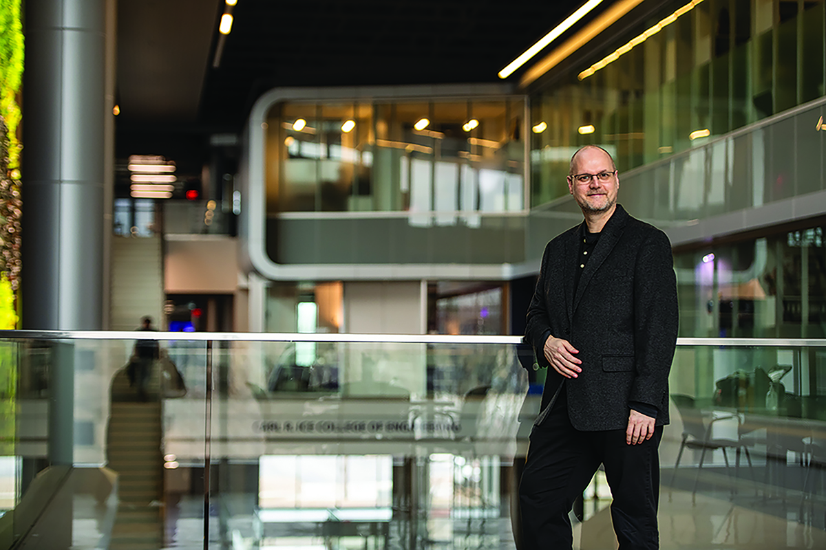 A man in all-dark professional attire stands in front of a glass divider with the background of the interior of a large building, including glass-walled conference rooms and a large staircase.