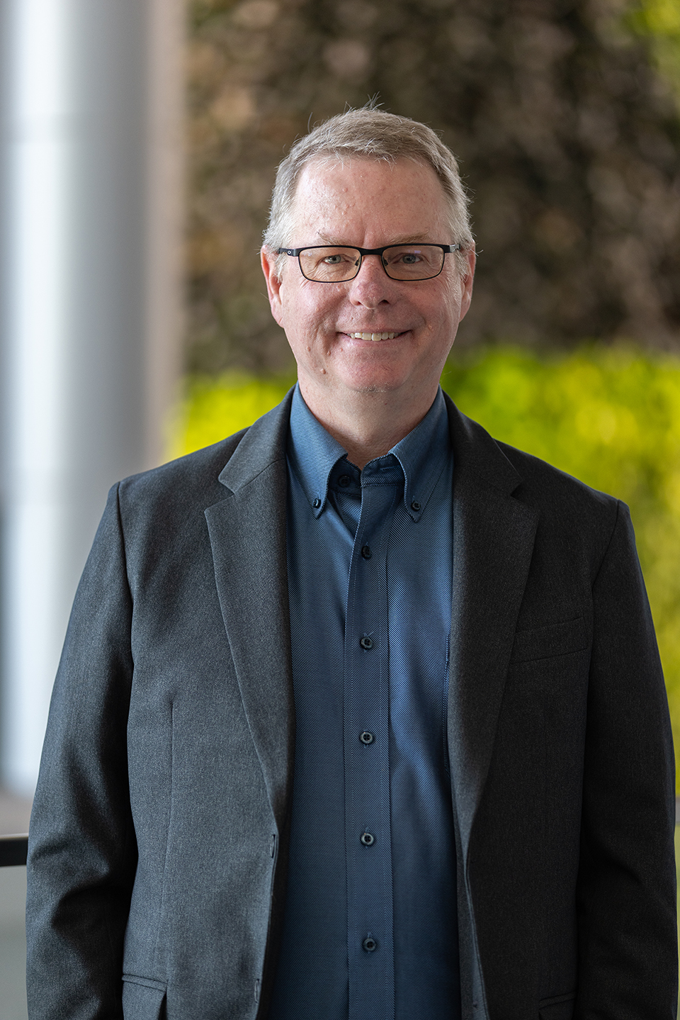 A man wearing a blue button-up collared shirt and a dark gray suit jacket smiles against a green wall background.