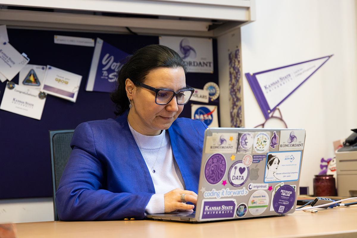 A woman wearing a dark purple blazer over a lavender sweater in thick dark glasses looks at a laptop that has many data and coding stickers on the back of it.