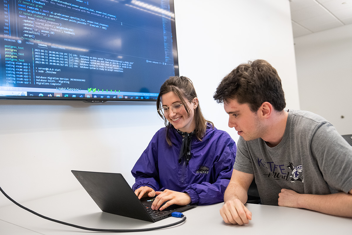 A female student in a purple windbreaker and a male student in a gray t-shirt look at a laptop screen that is projected on a TV behind them, which shows code.