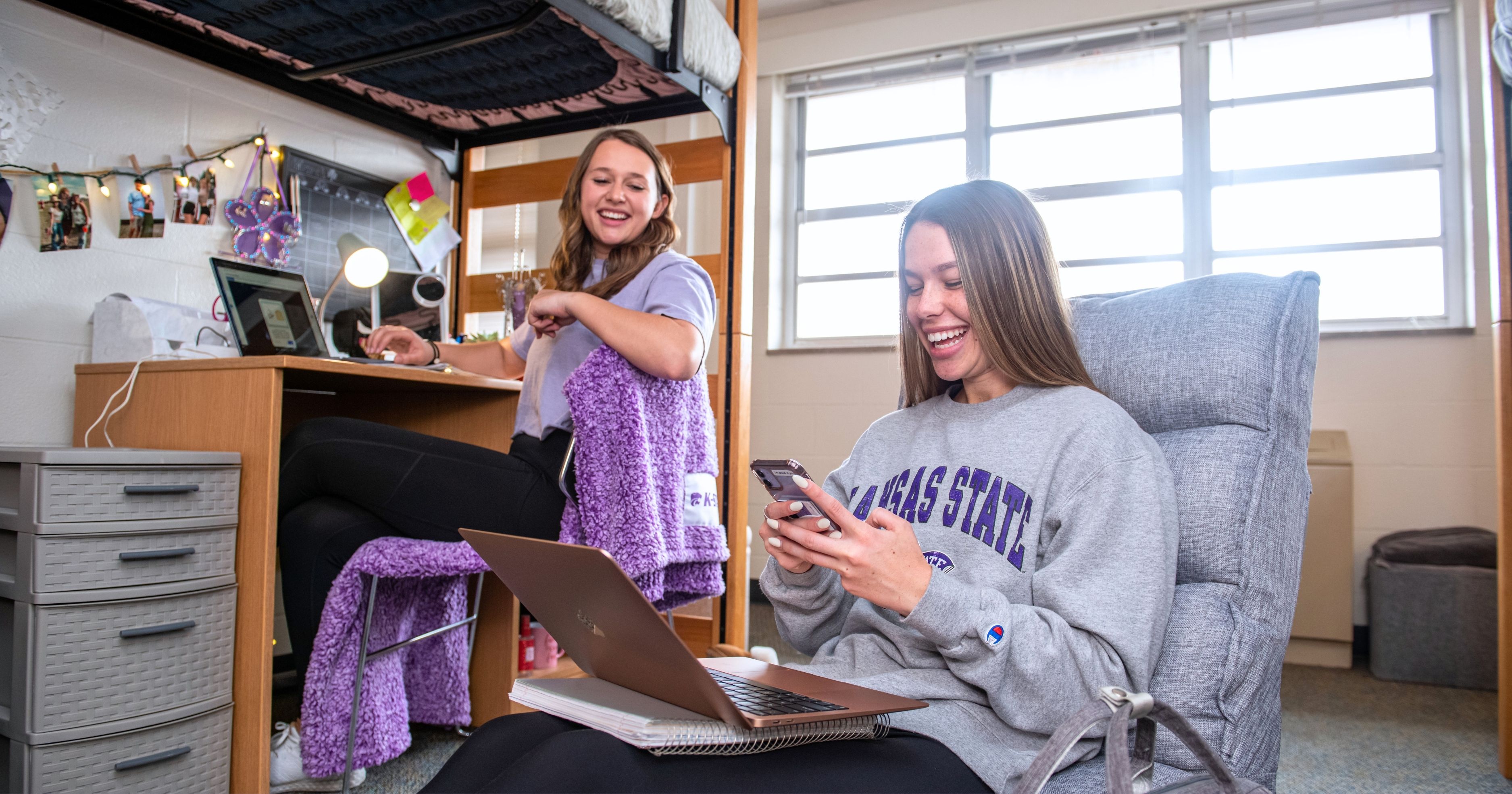 Two students sit in a dorm room. One sits on a beanbag chair and smiles down at a phone. In the background, another student sits at a desk.
