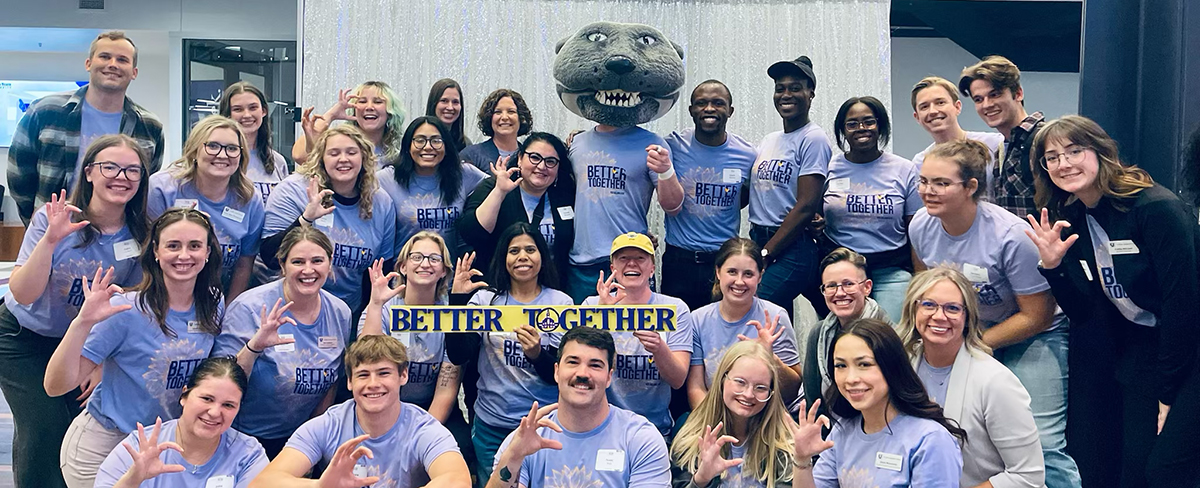 A large group of people wearing purple shirts surround mascot Willie the Wildcat. A student holds a sign that says "Better together."