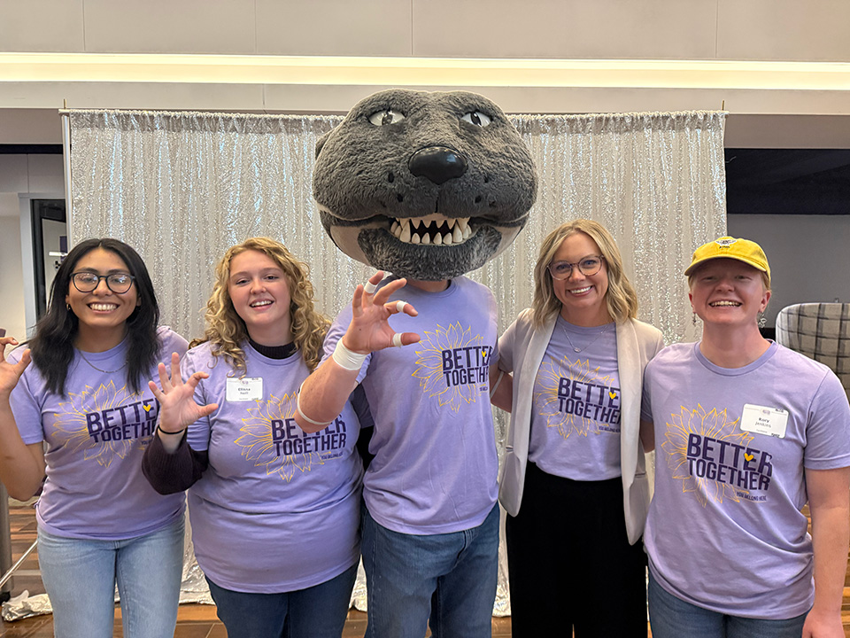 Four people stand with Willie the Wildcat, K-State's mascot, wearing matching purple shirts.
