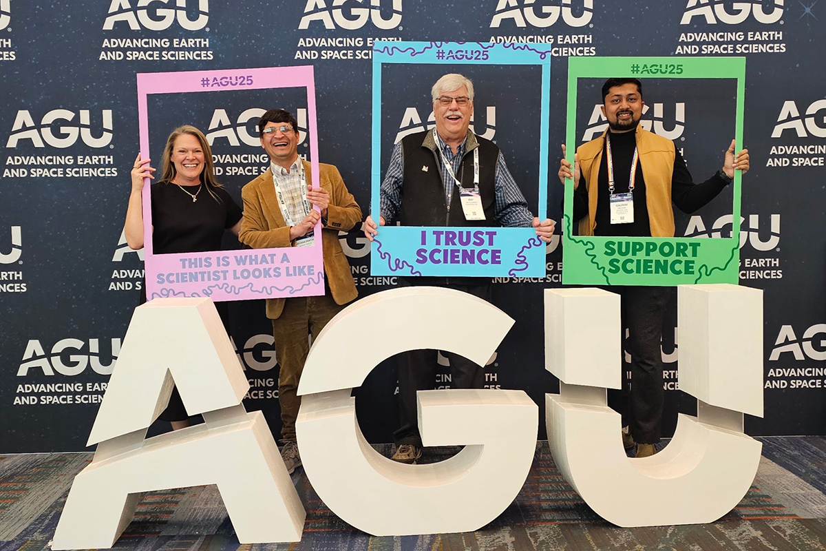 A group of college researchers pose for a portrait behind big block letters that spell A-G-U in a conference room in front of a backdrop.