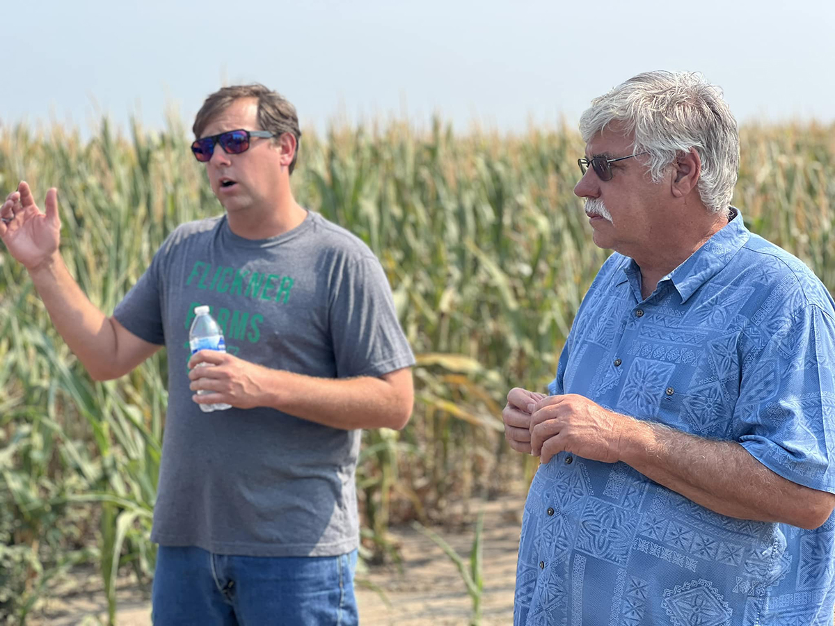 An adult man and his father stand and present about their farm in front of a corn field.