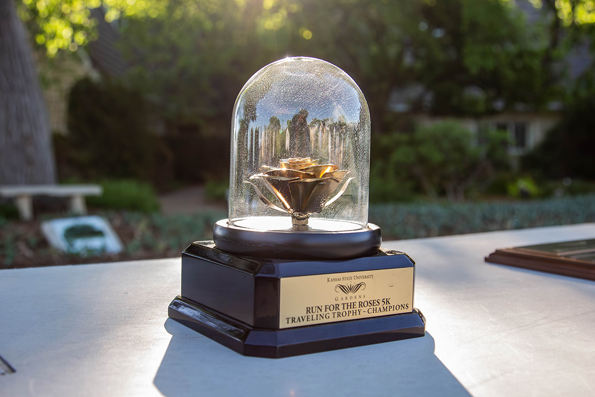 A golden rose trophy encased in a glass dome sits on a wooden base engraved "Kansas State University Run for the Roses 5K Traveling Trophy - Champions" on an outdoor table with a blurred park background.