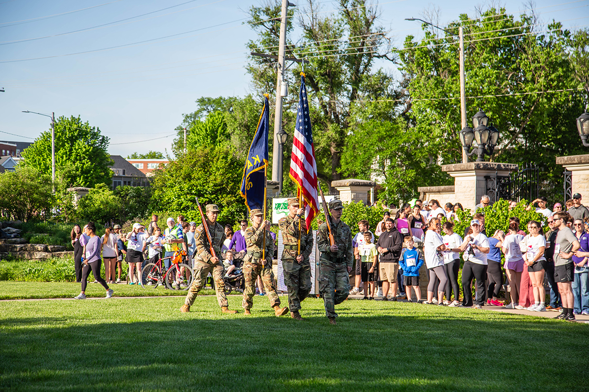 Kansas State University Army ROTC cadets in camouflage uniforms present the American flag and military colors on a grass field as a large crowd of Run for the Roses 5K participants stands at attention in the background.