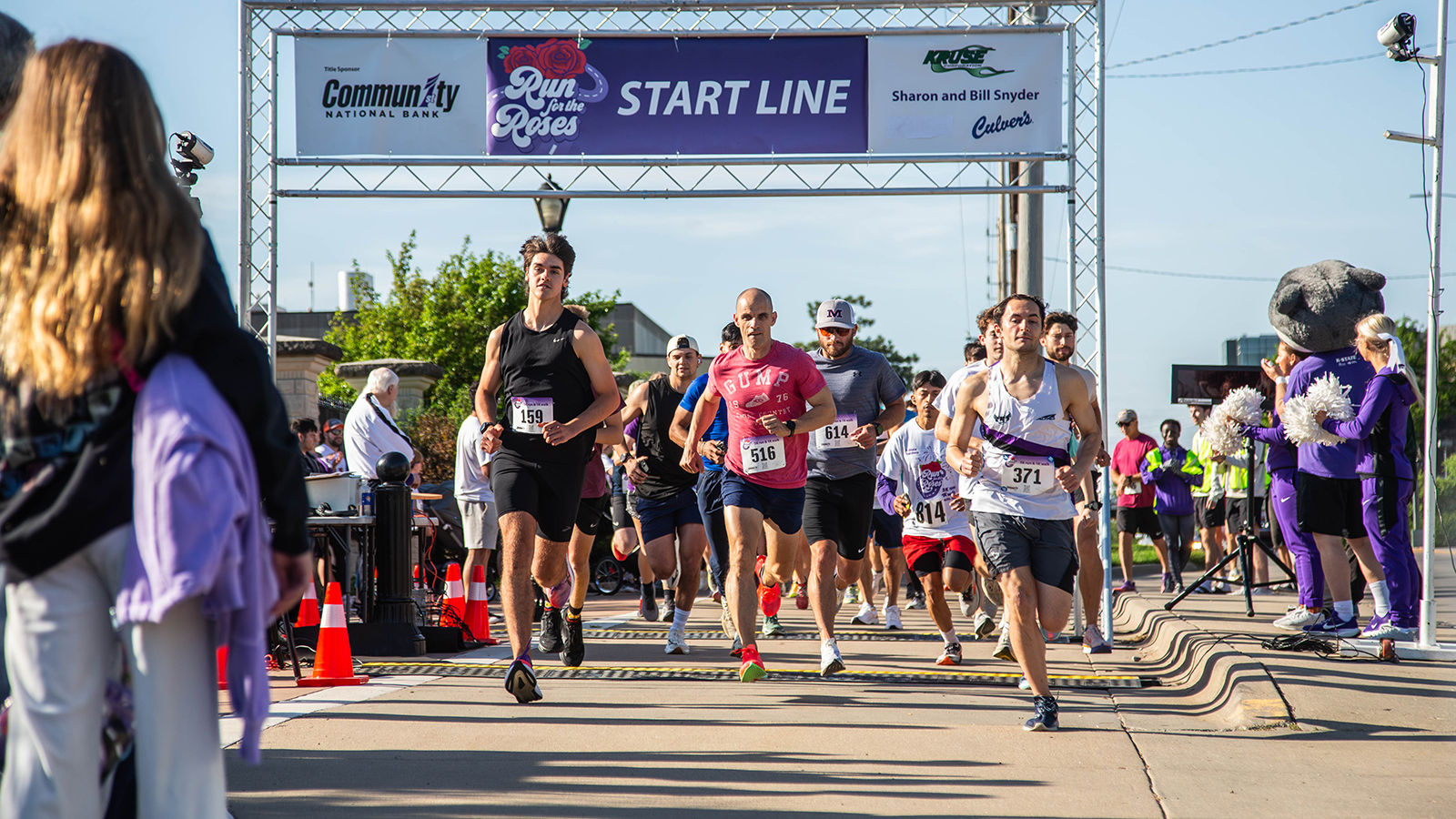 A group of runners run toward the camera from under a race starting tower on a sunny day on a college campus.