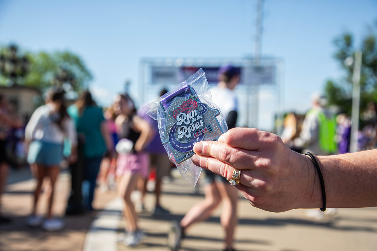 A hand holds a commemorative Run for the Roses 5K pin in a clear plastic bag with the event logo featuring a rose design as participants and the start line banner appear blurred in the background.