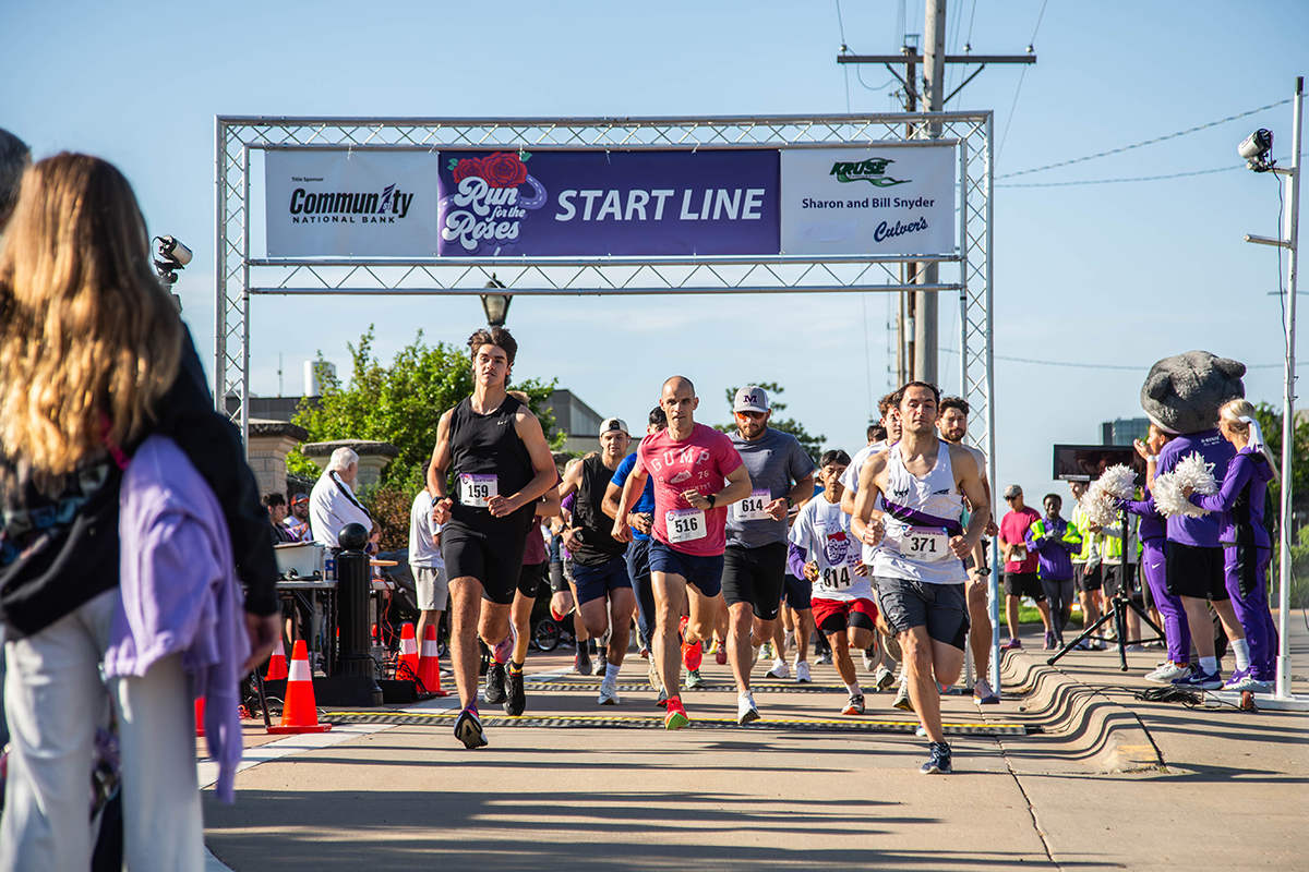 A group of runners run toward the camera from under a race starting tower on a sunny day on a college campus.