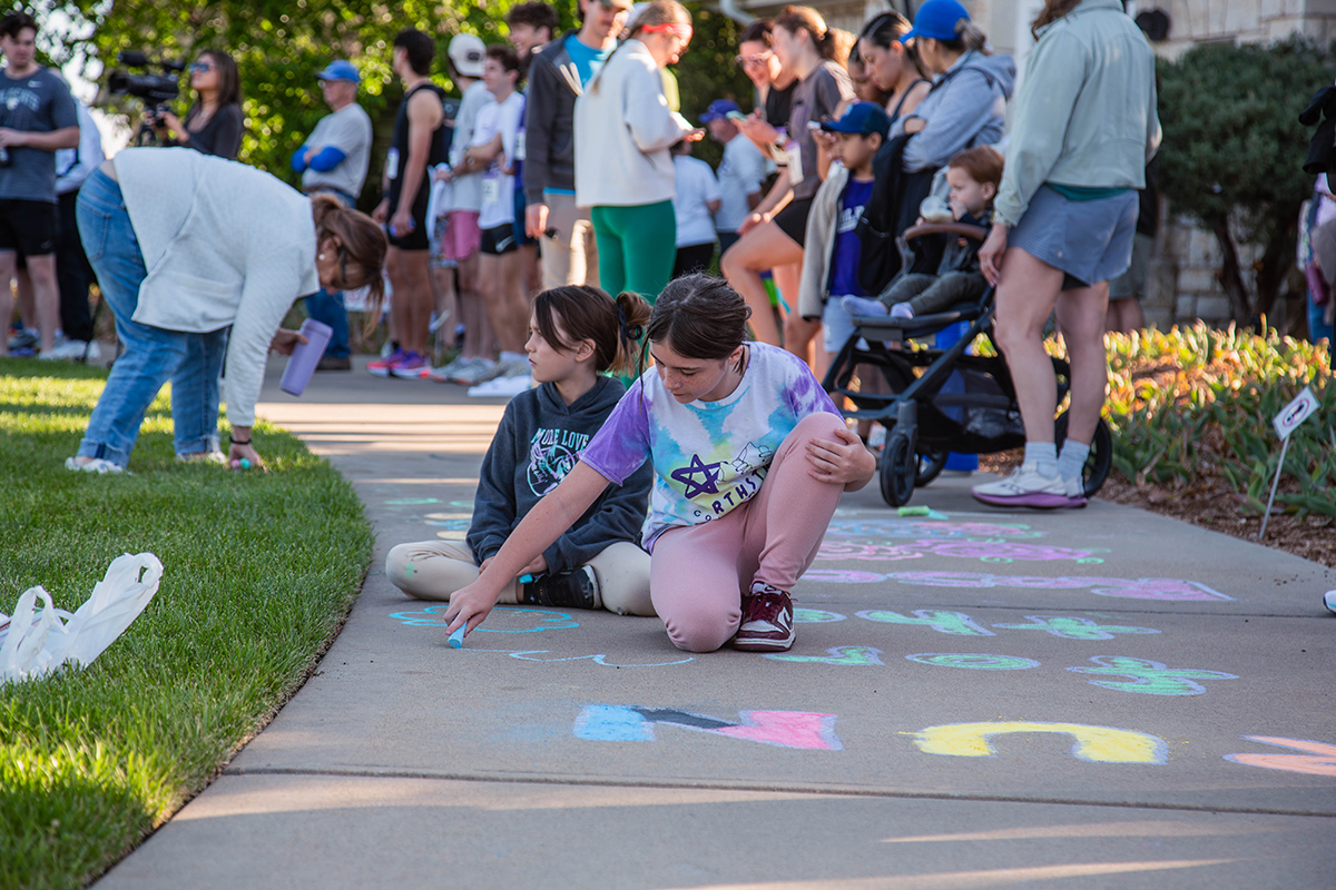 Two girls sit on a sidewalk drawing with colorful chalk while families and spectators gather in the background.