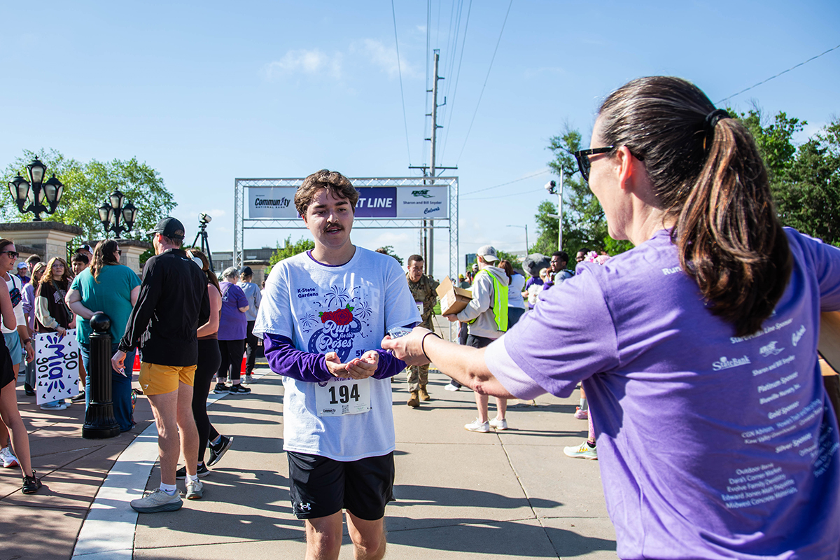 A male runner wearing race bib number 194 and a Run for the Roses shirt receives a medal from a volunteer in a purple event shirt near the start line as spectators gather in the background.