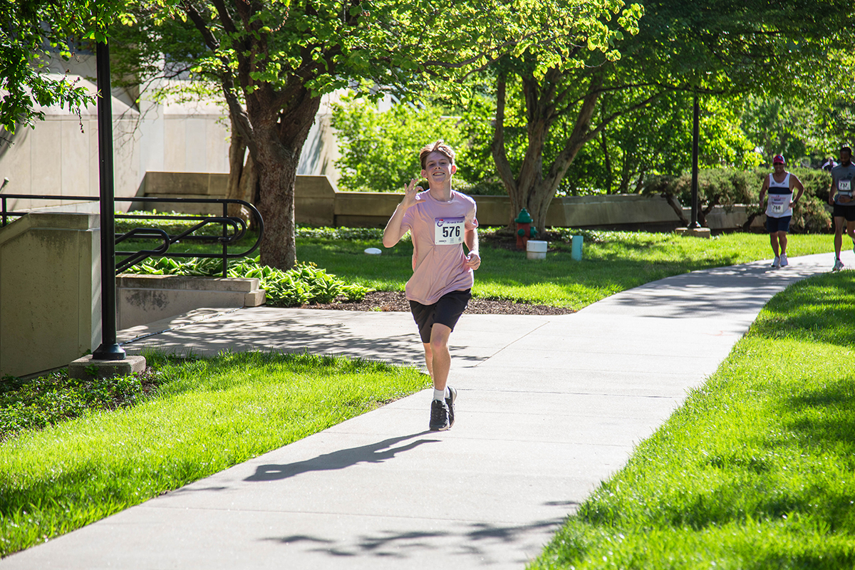 A young male runner wearing a pink shirt, visor and race bib number 576 runs along a tree-lined sidewalk through a sunlit campus park during the Run for the Roses 5K as other runners follow behind.