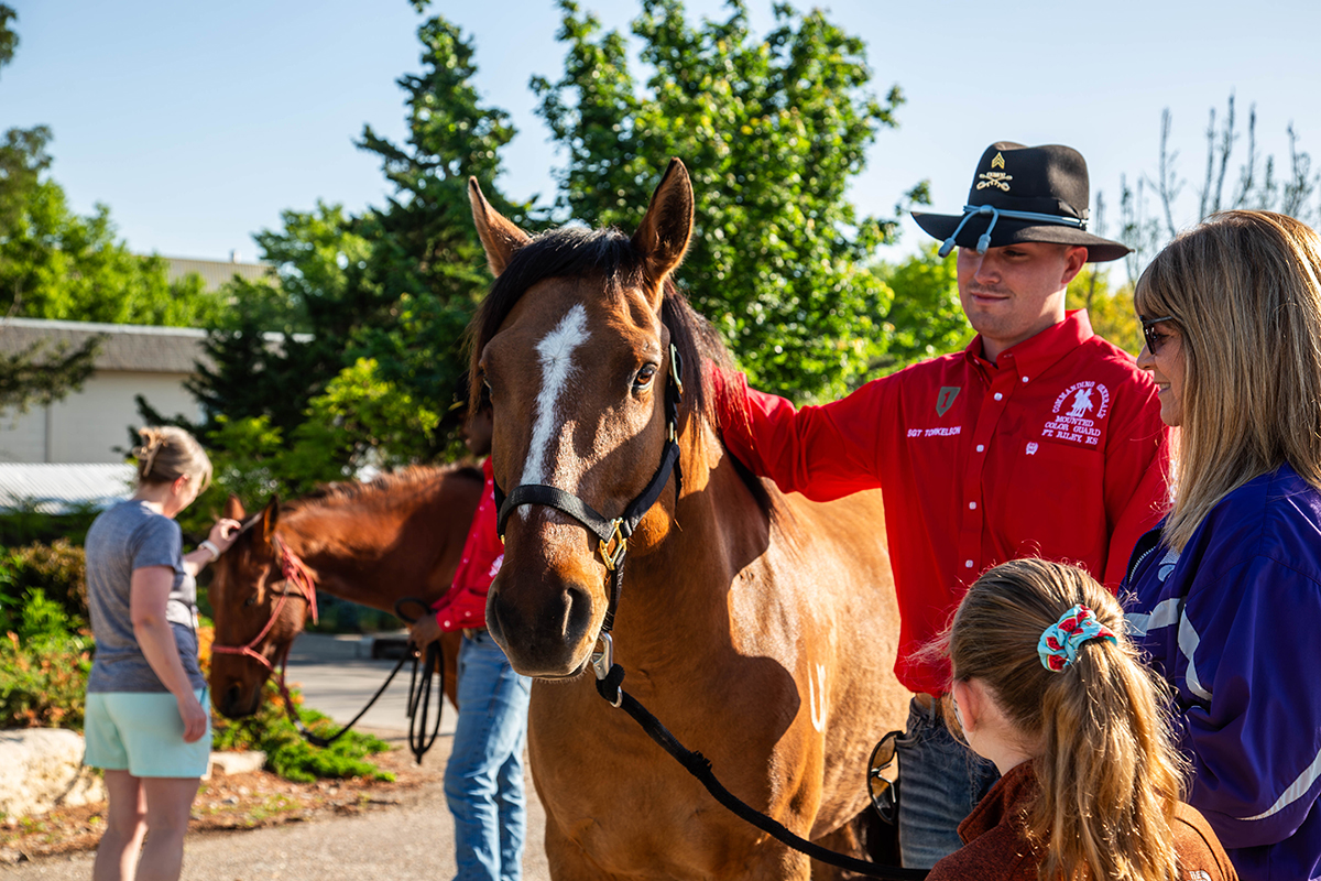 A Fort Riley Color Guard member in a red shirt and black cowboy hat pets a brown horse while two young girls look at the horse. 