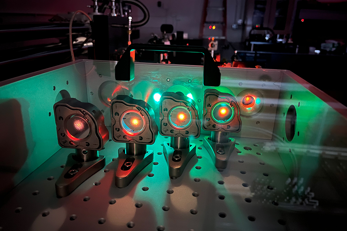 A row of optical instruments on a lab bench emit red and green laser light, illuminating mirrors and components used in a physics experiment.