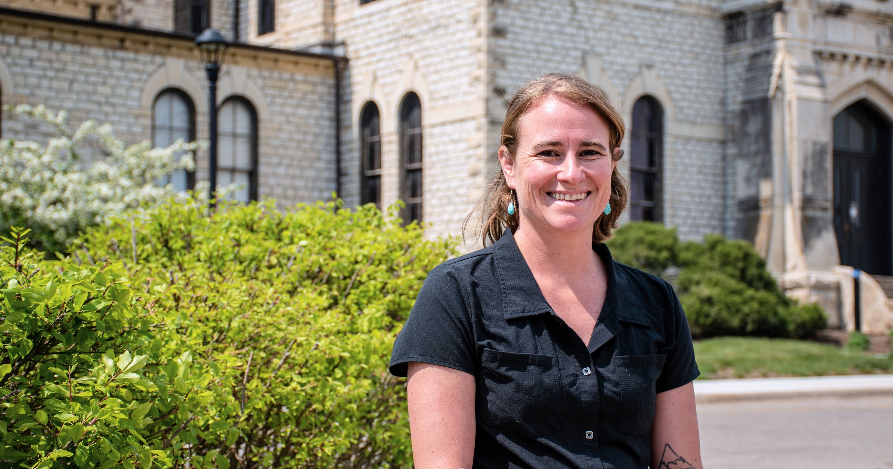 Morgan Greene smiles for a photo in front of Anderson Hall. She wears a black short-sleeve button-up shirt and turquoise earrings. A tattoo of a mountain is visible on her forearm.