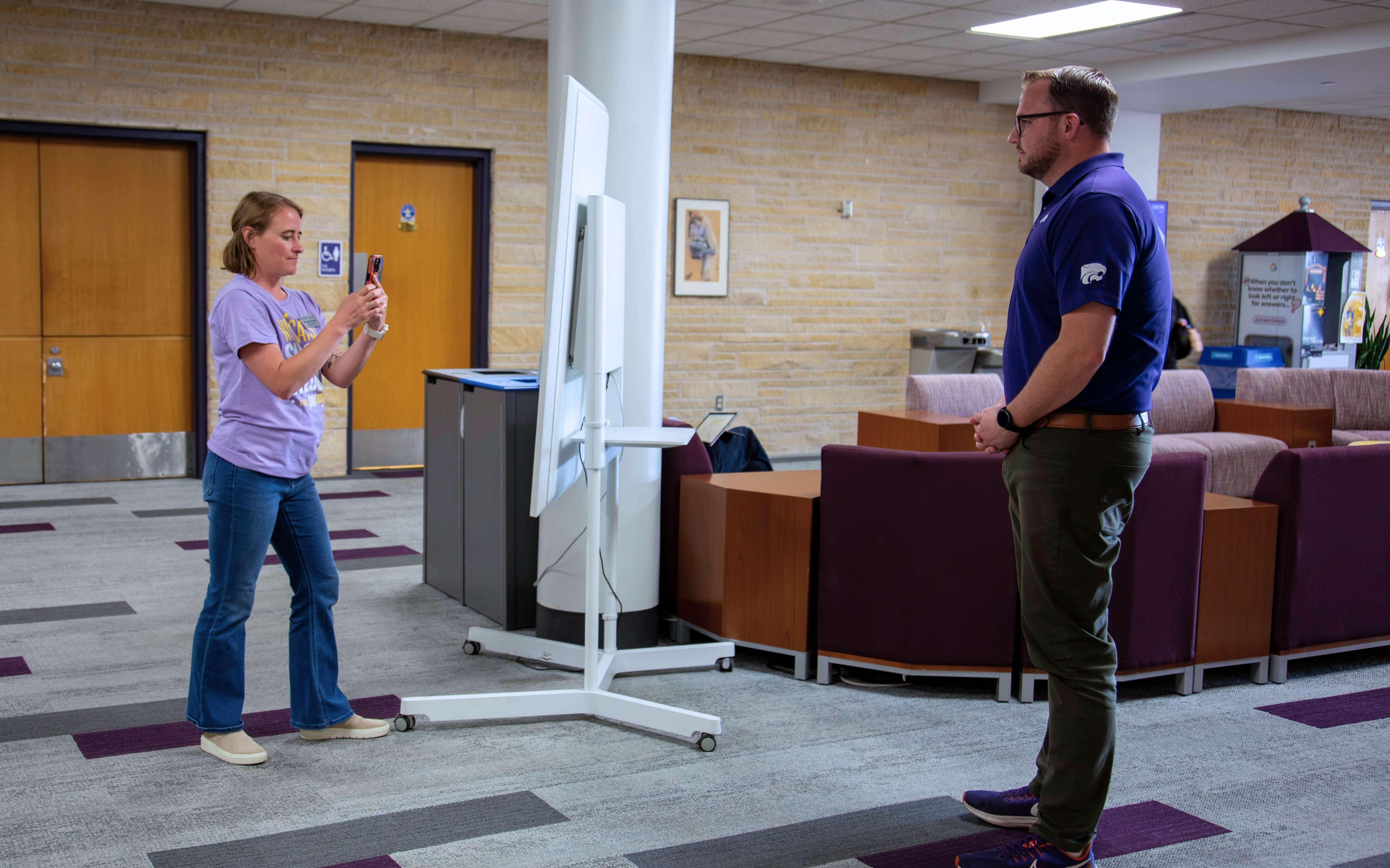 Morgan Greene, wearing jeans and a lavendar t-shirt, holds a phone to take a video of a man in a purple K-State polo. They both stand in an open seating area in the K-State Student Union.