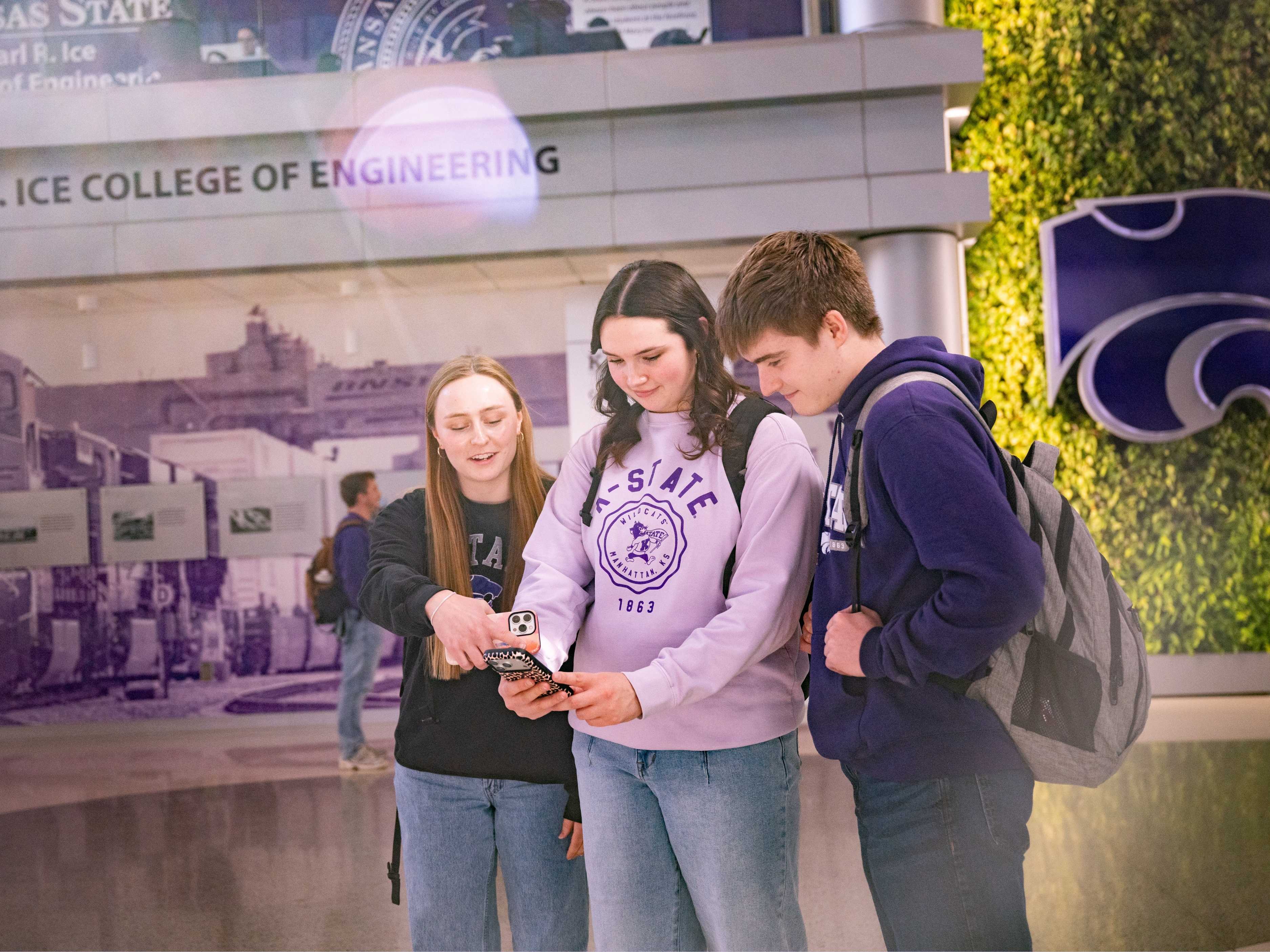 Two female and one male engineering student stand looking at a phone screen. They all wear K-State sweatshirts. In the background is a wall with fake greenery and a purple powercat.