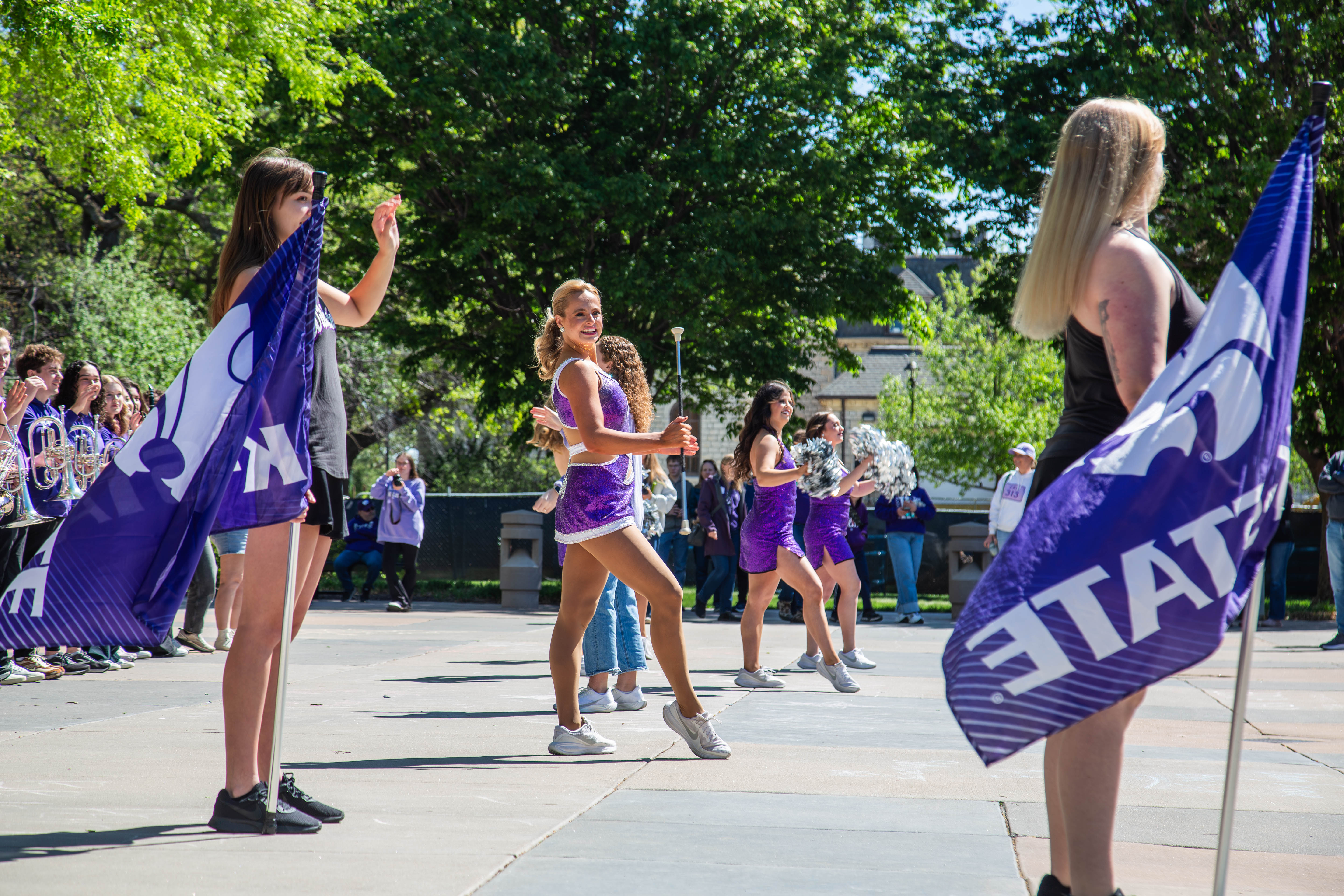 K-State twirlers and spirit squad perform outdoors on campus, waving purple flags as a crowd and band members look on. 
