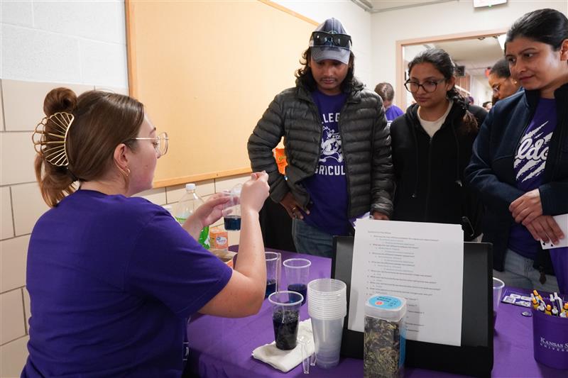 Three Kansas State students in purple shirts and blue gloves pose with large bags of popcorn in a food processing facility on campus. 