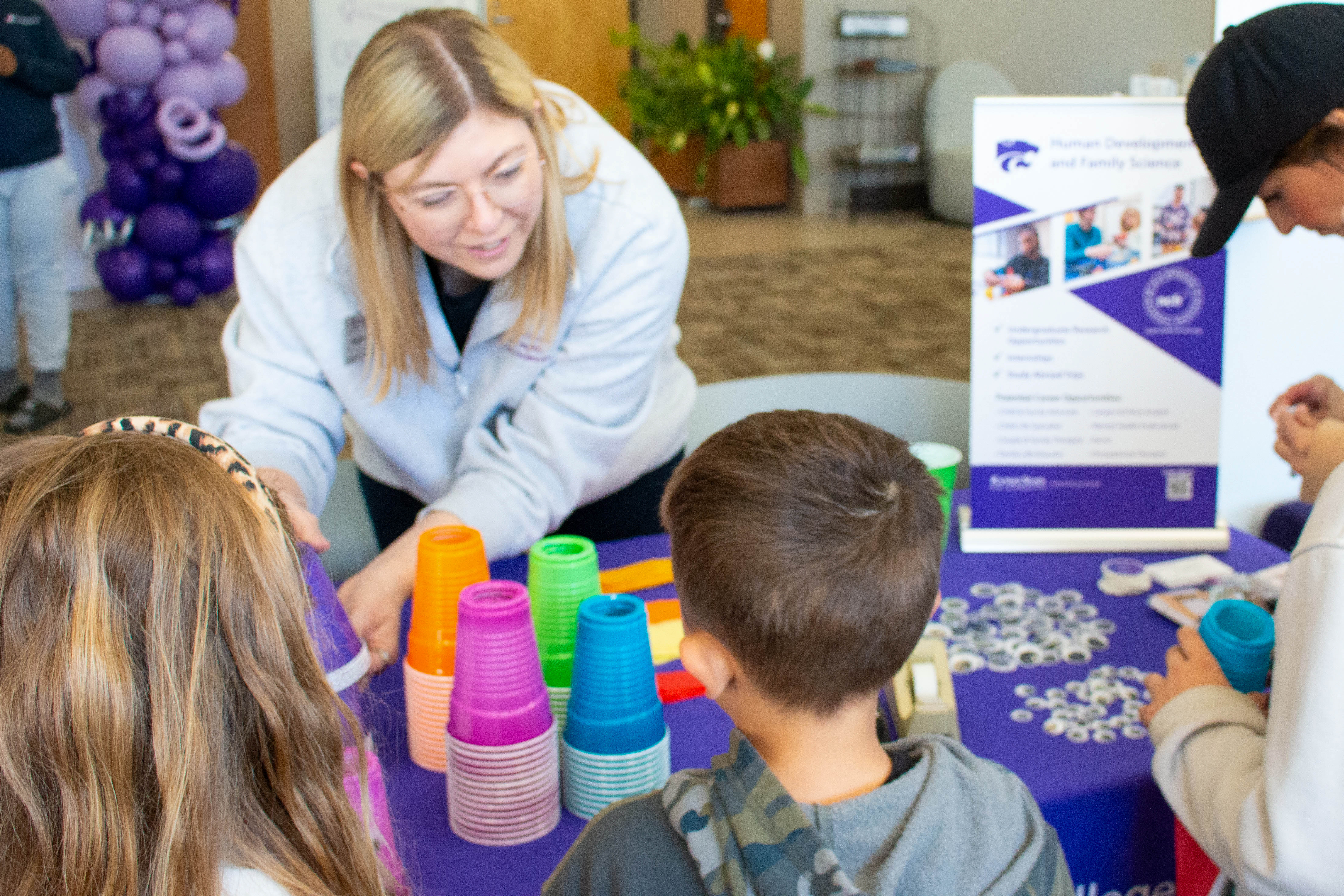 Kansas State Human Development and Family Science students engage children in a colorful cup stacking activity at a purple-decorated table with K-State Wildcats promotional materials. 