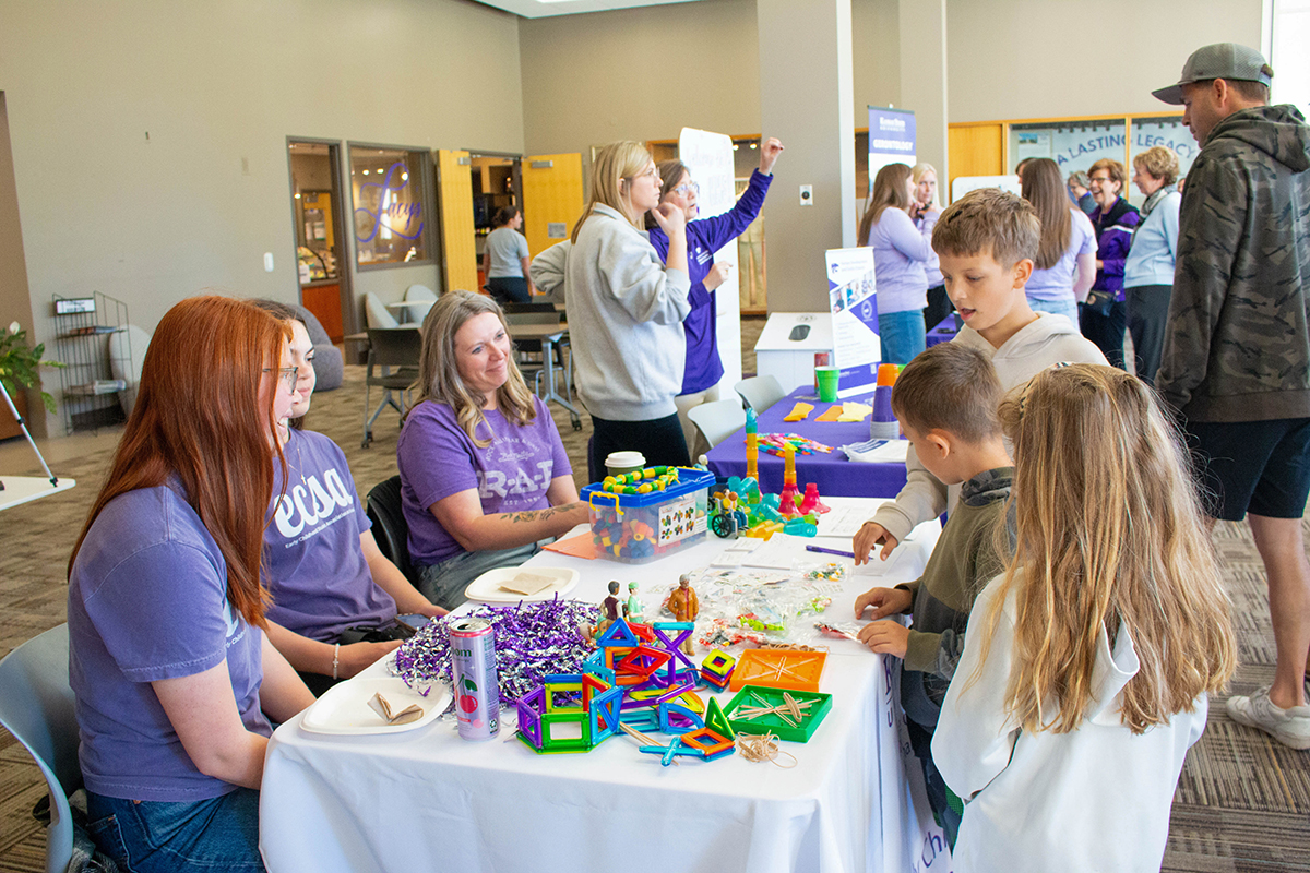 Kansas State students in purple shirts engage children with hands-on building activities using colorful blocks and magnetic tiles at an interactive table during a campus outreach event. 