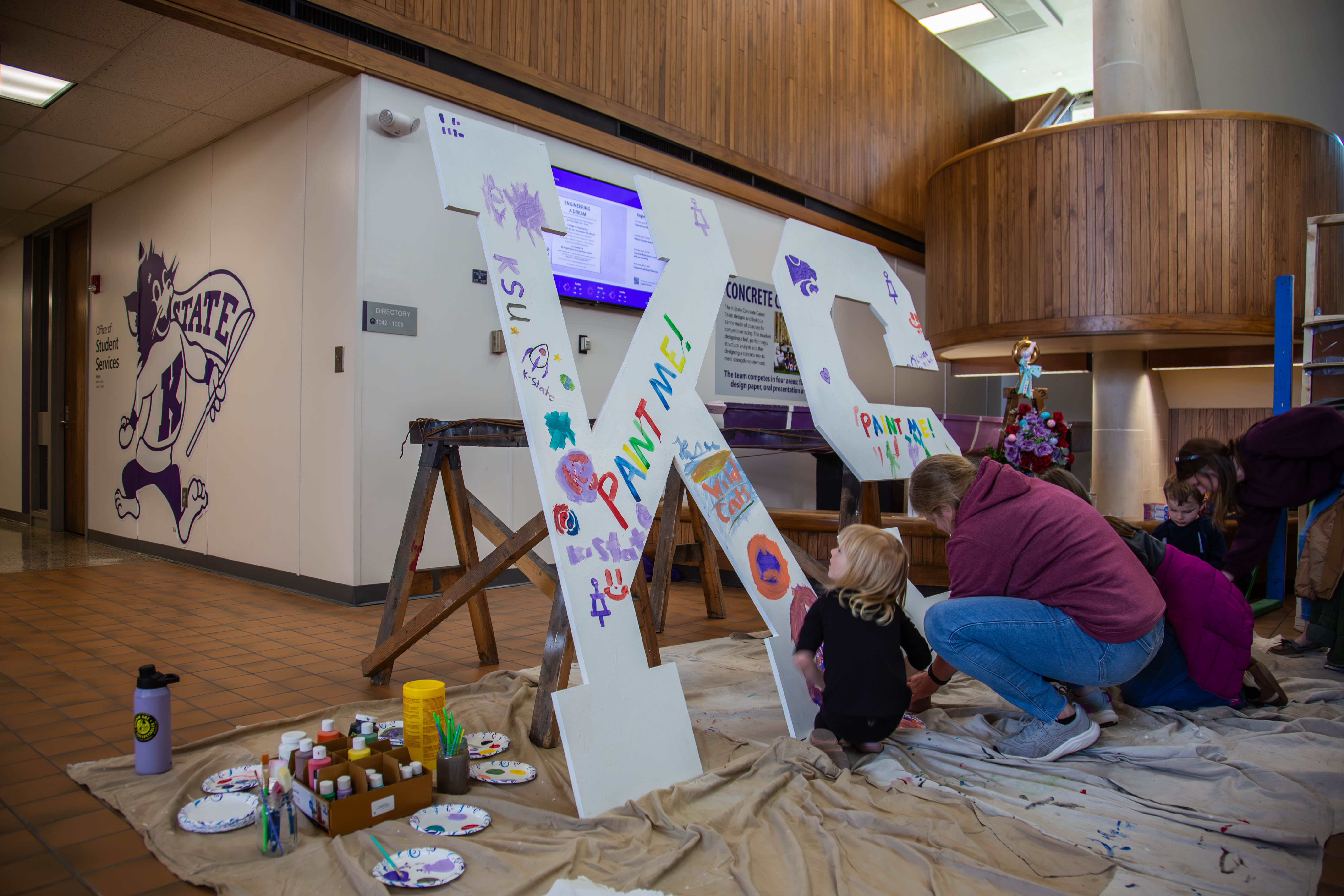 Families paint colorful designs on large K-State letters in the Kansas State Student Union lobby, with the Wildcats mascot logo visible on the wall behind them. 