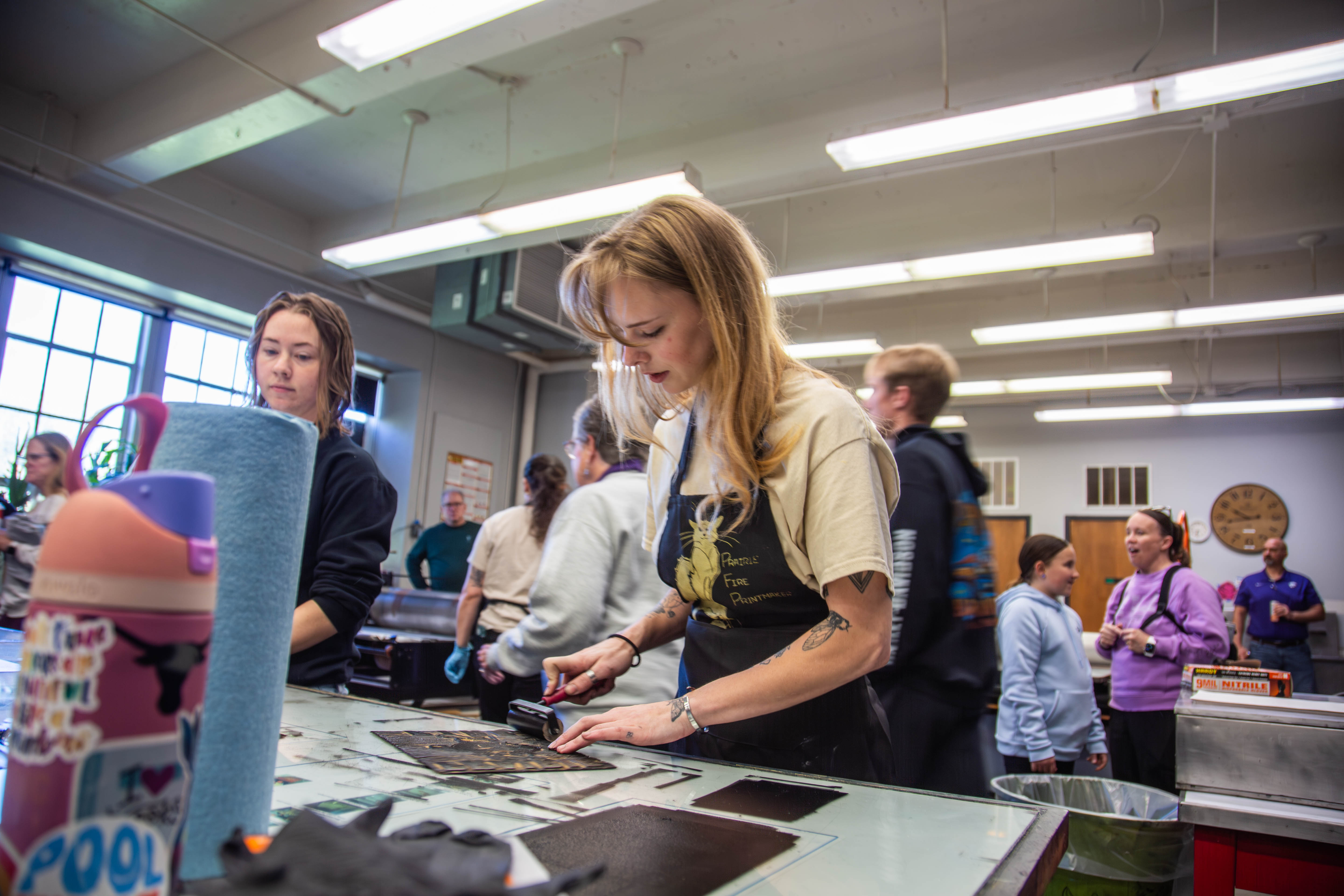 A Kansas State student in an apron demonstrates printmaking techniques using a brayer on paper in a campus art studio while children and families observe during an outreach event.