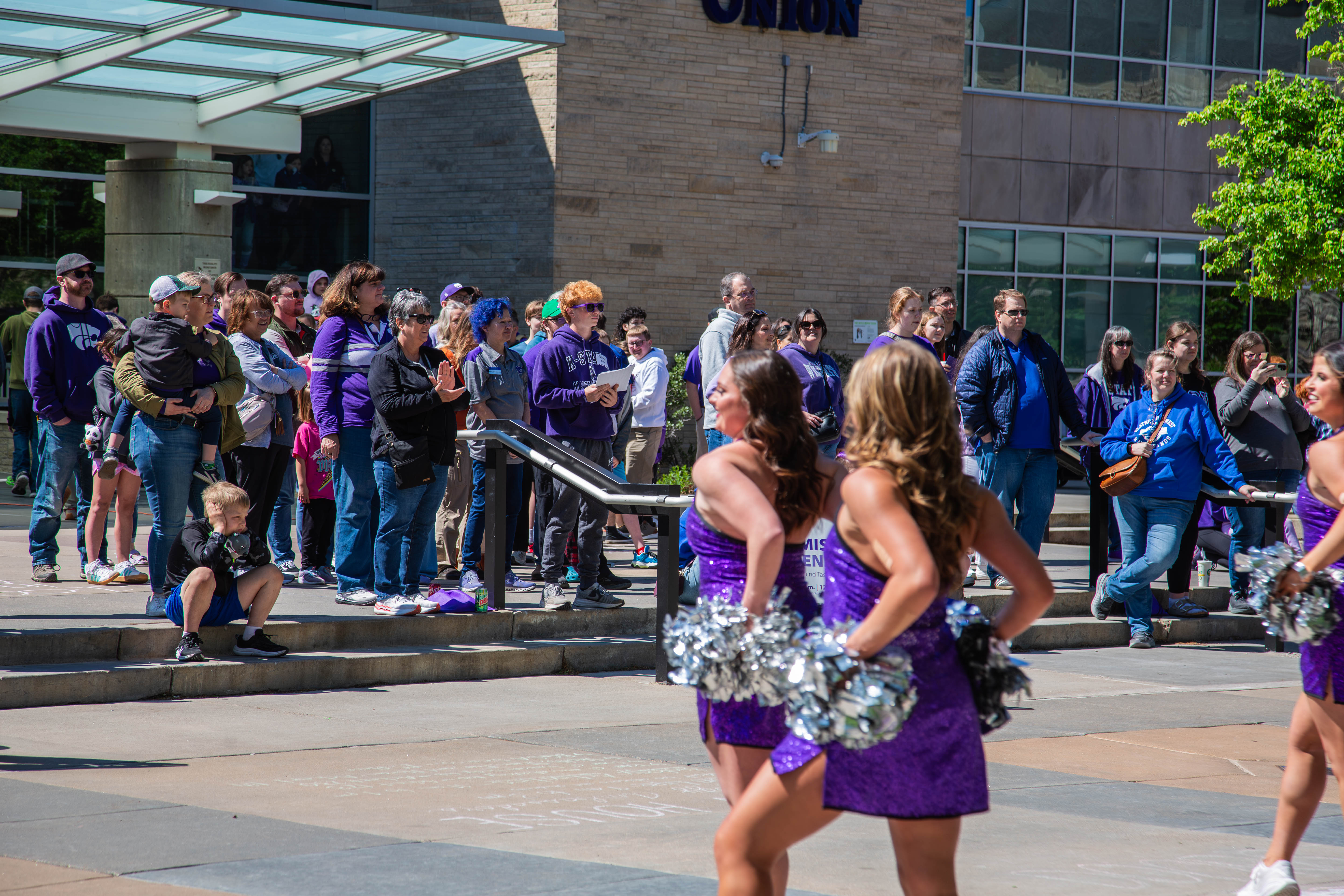 Kansas State cheerleaders in purple uniforms with silver pom-poms perform during a campus parade as spectators in K-State purple line the steps of a brick building. 