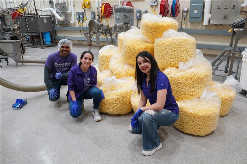 Three Kansas State students in purple shirts and blue gloves pose with large bags of popcorn in a food processing facility on campus.