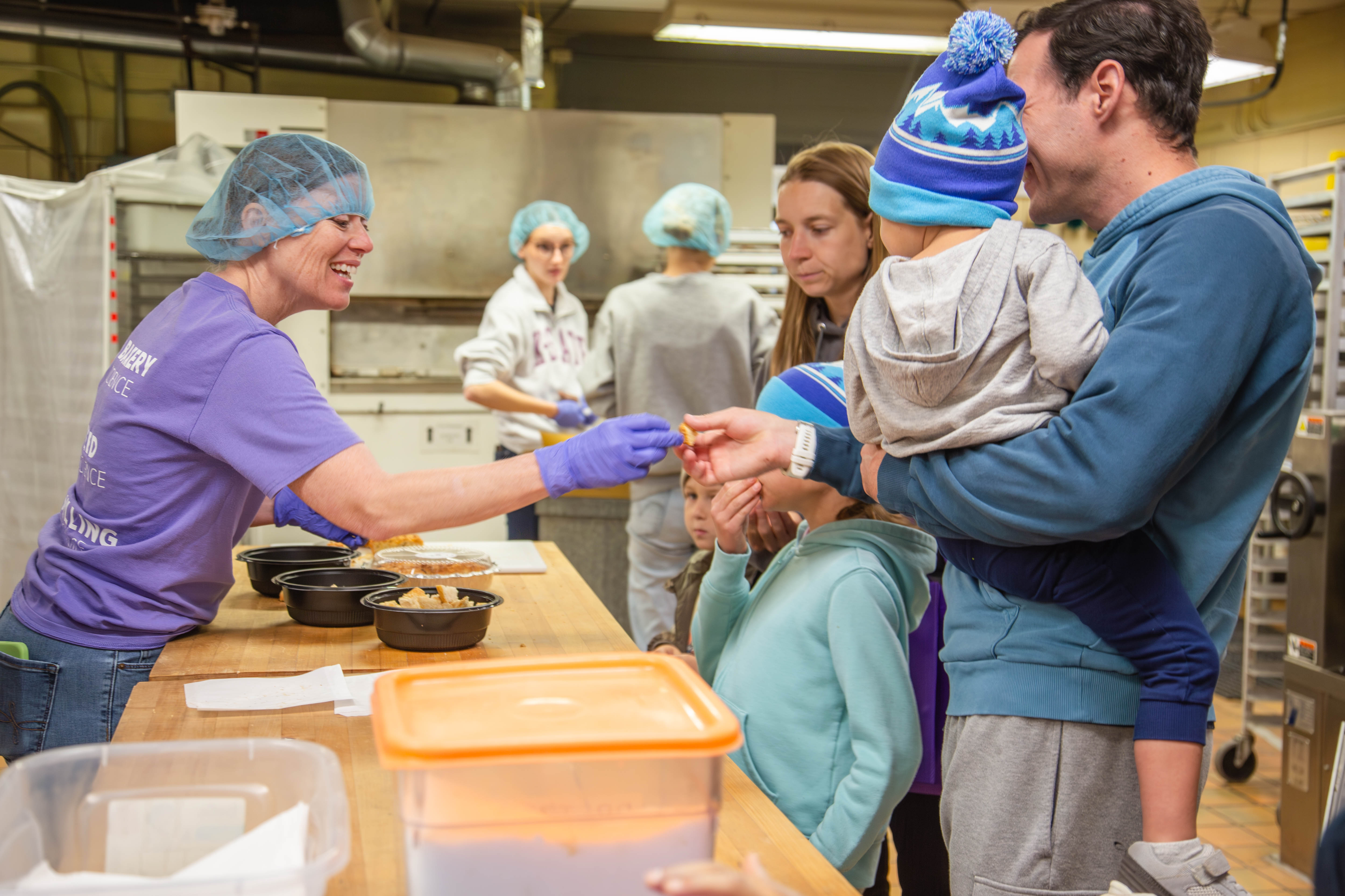 A Kansas State food science instructor in a purple shirt and hairnet offers samples to a family with young children during a campus food production demonstration. 
