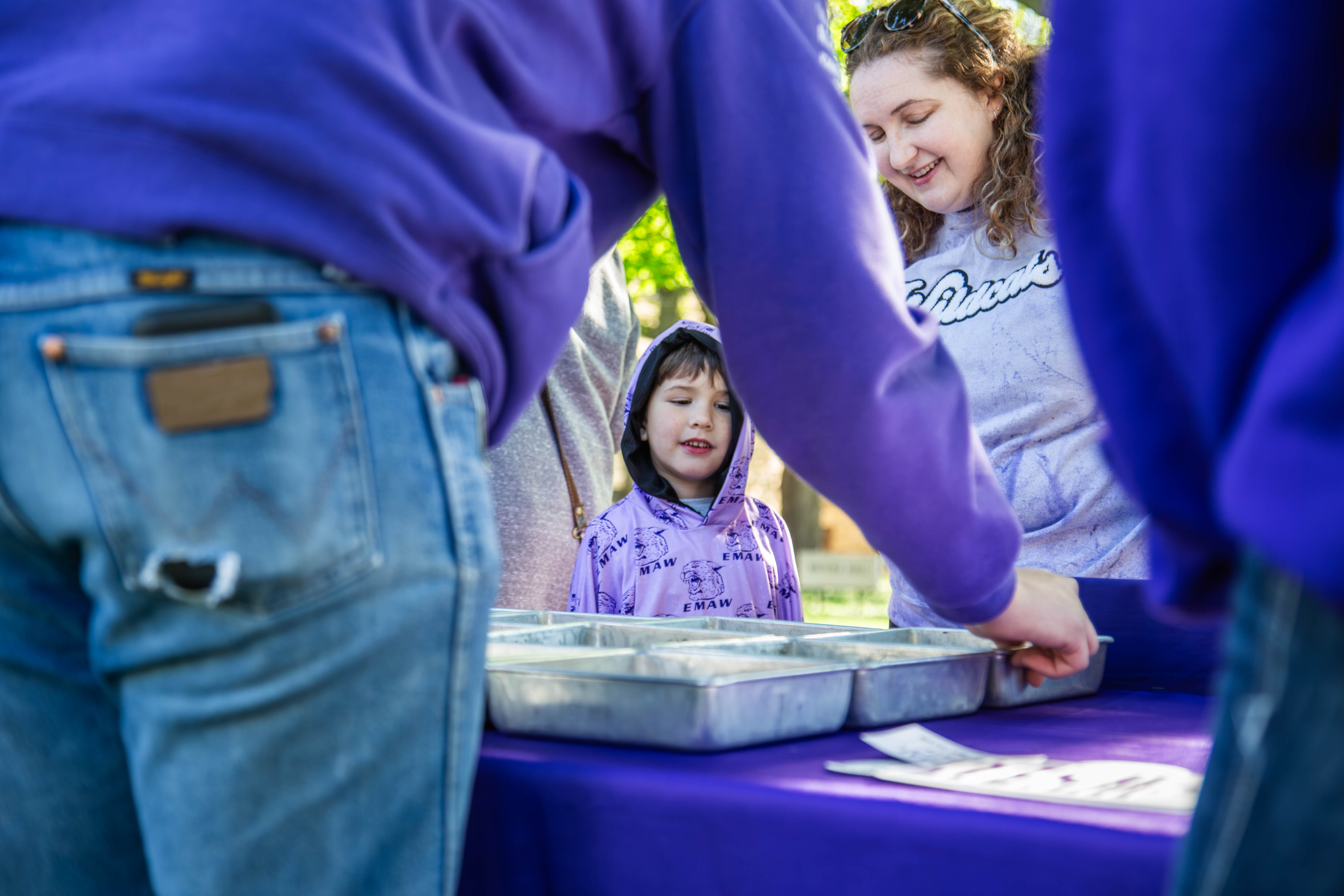 A Kansas State volunteer in a purple shirt presents a food tray to a smiling child in a purple hoodie and parent at an outdoor campus community event. 