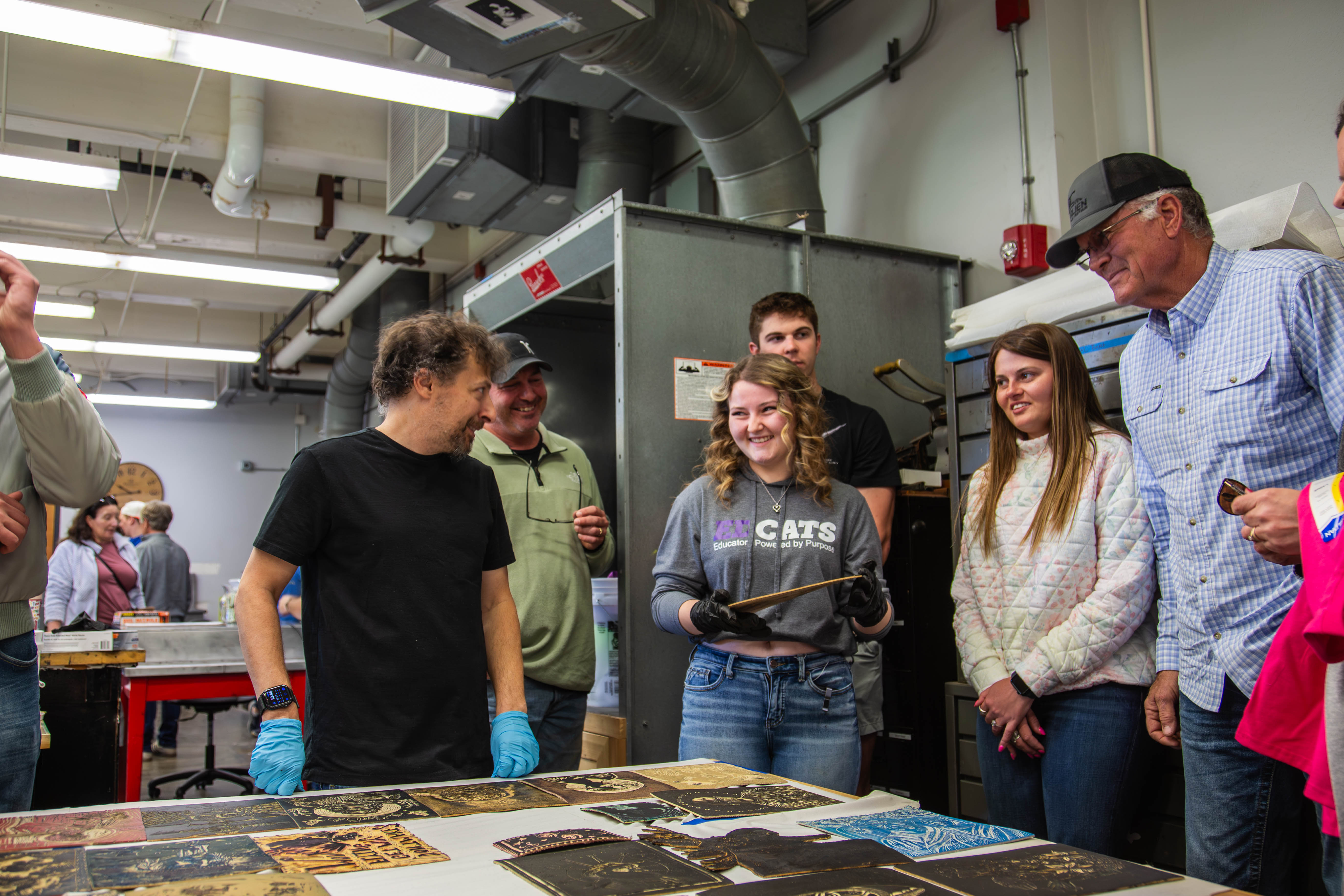 A Kansas State printmaking student demonstrates techniques to visiting families gathered around a table displaying various prints in a campus art studio. 