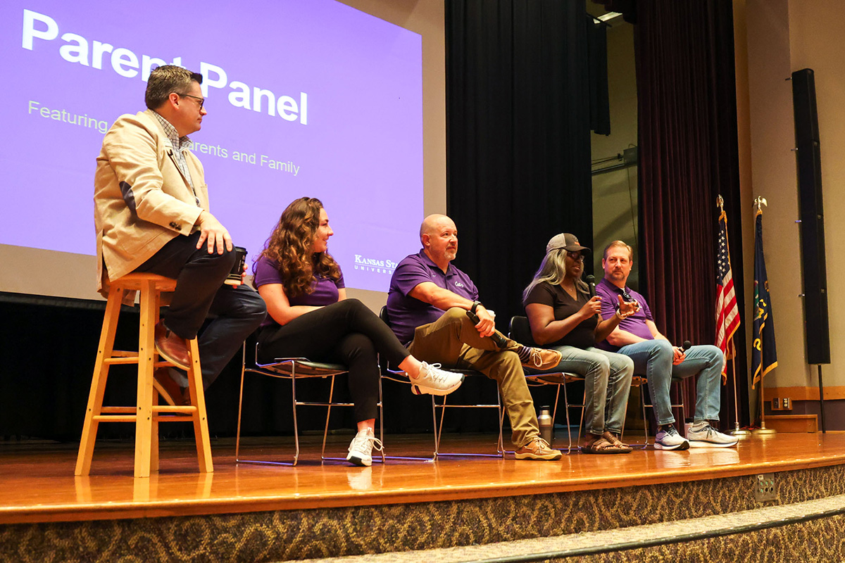 A Kansas State University staff member facilitates a parent panel during a new student orientation. A group of parents sits on a stage, listening to one of them speak to the audience.