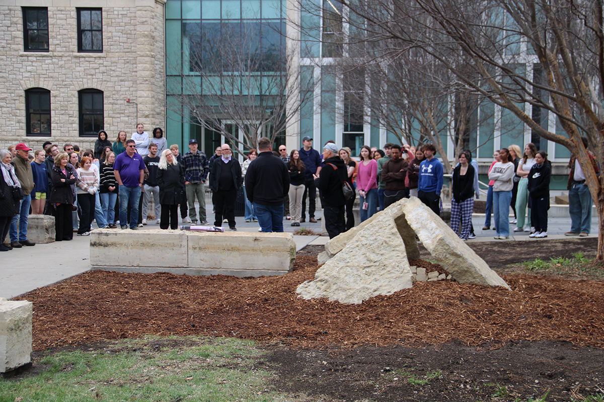 A group of college students, faculty and staff gather for the dedication of a limestone sculpture in front of a glass-walled academic building on a college campus.