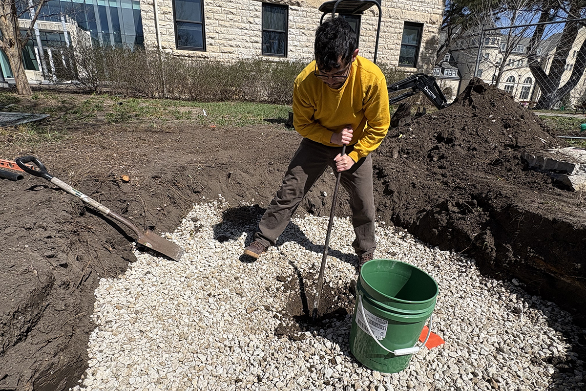 A man in a yellow-long sleeve shirt uses a compacting tool to stamp down pieces of limestone gravel at the bottom of a shallow dirt pit on a college campus's grounds.