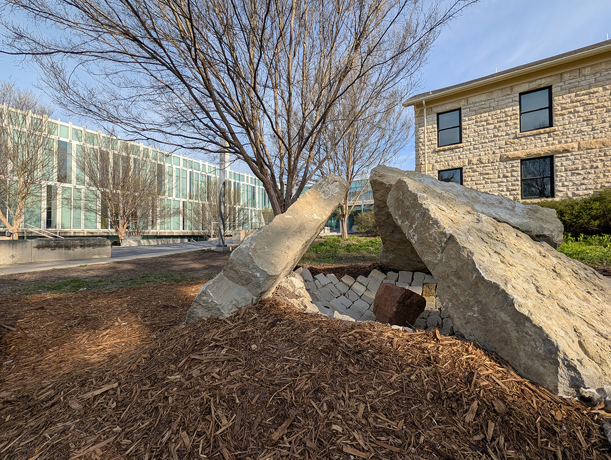 Three large slabs of limestone overlooking a rounded hunk of red clay brick form an outdoor sculpture on a college campus.