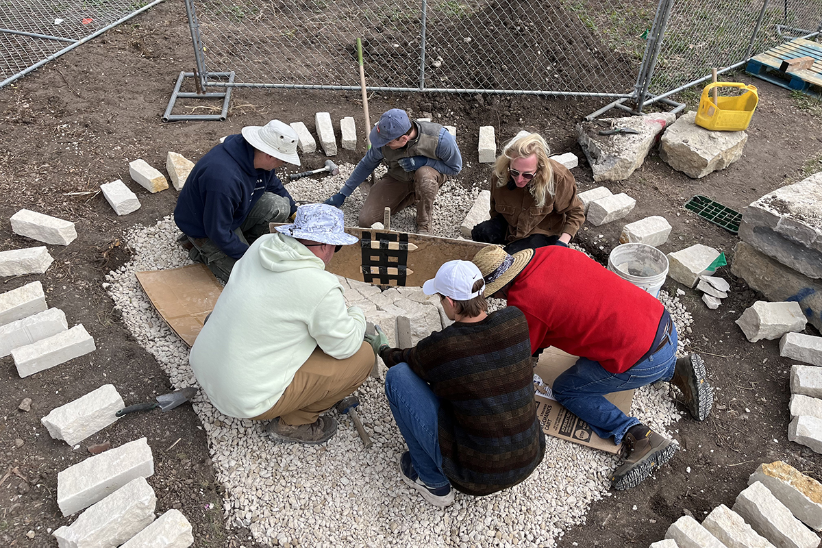A group of college students in cold-weather clothing sit on top of a bed of gravel in a shallow dirt pit and arrange large limestone bricks into a rounded grid on top of it.