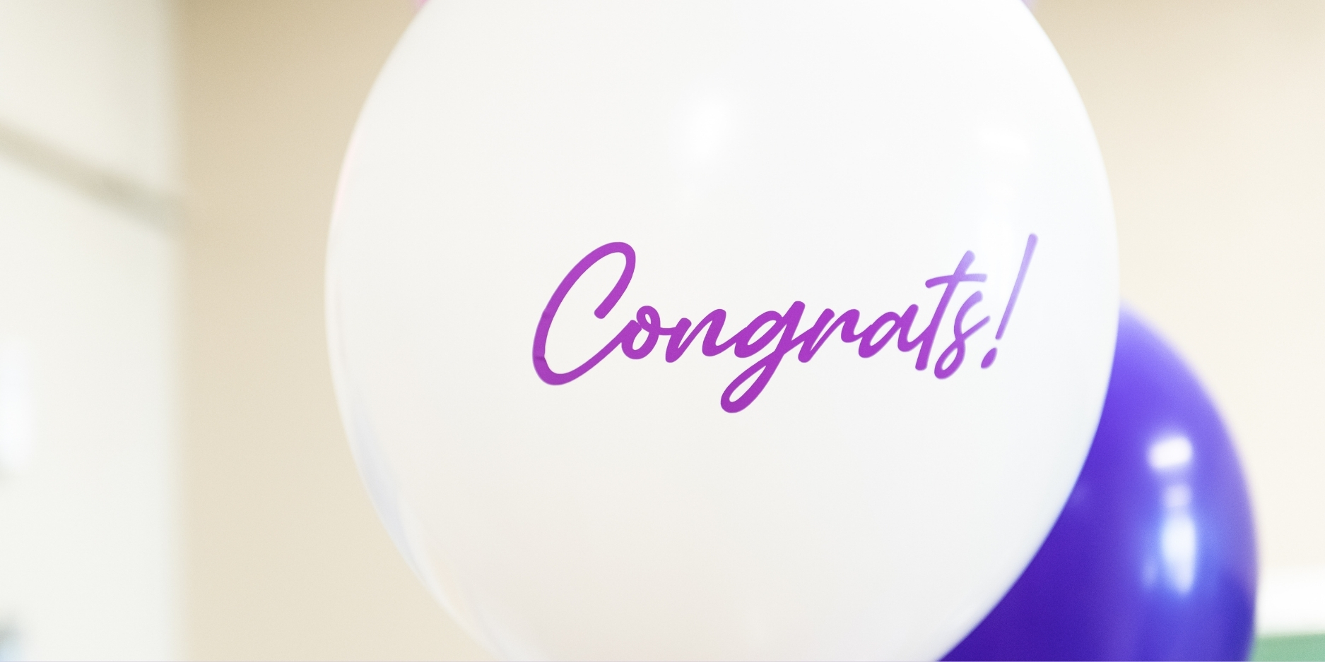 A close up photo of a white balloon with purple script font spelling "Congrats!"