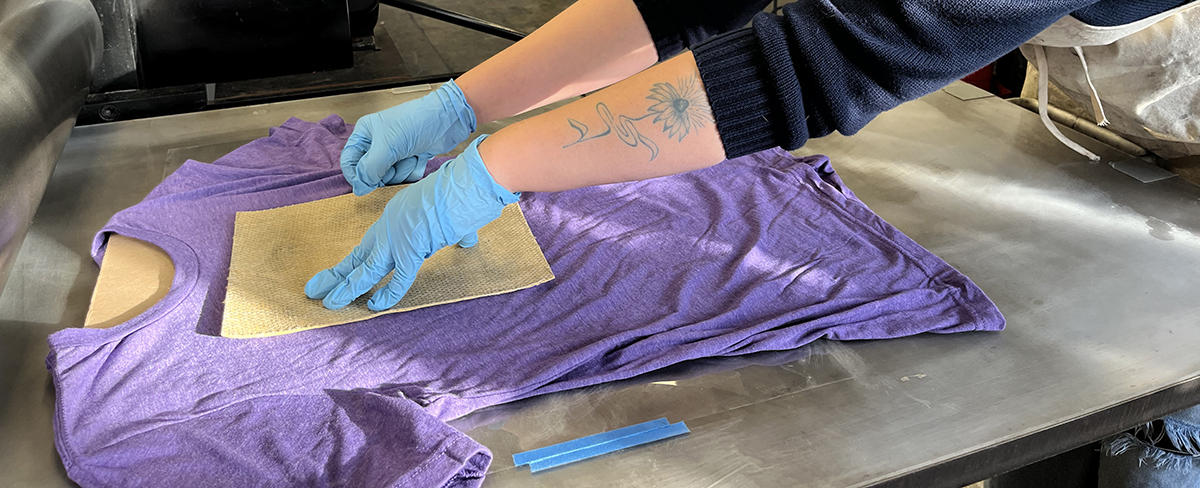 Two arms can be seen, one with a flower tattoo, pressing a beige piece of fabric onto a blank purple t-shirt.