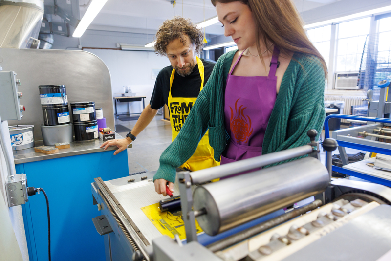 A student with long brown hair wearing a green cardigan over a purple apron works with printing material while a professor watches.