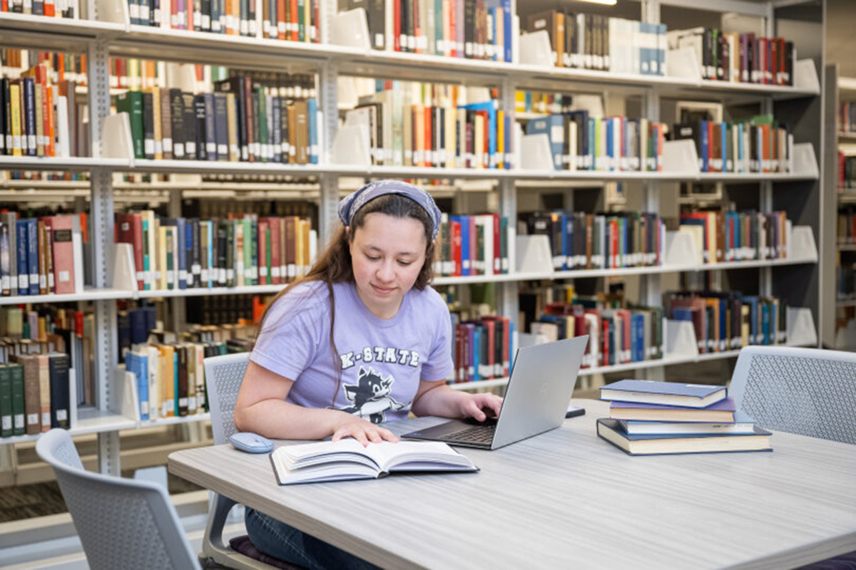 Student sits at a table in a library, reading an open book while using a laptop, with stacks of books nearby and shelves of books in the background.