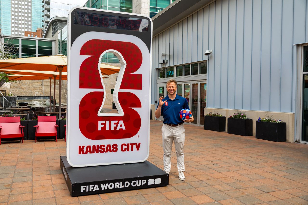 Ben Tenbrink stands next to a large outdoor FIFA KC sign while holding a soccer ball. 
