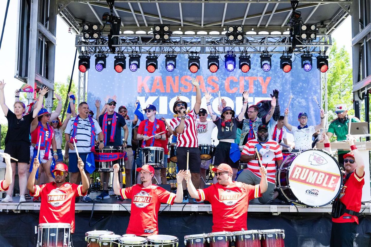 An outdoor stage with red and blue lights contains many musicians and entertainers celebrating. A banner behind them says Kansas City Kickoff.