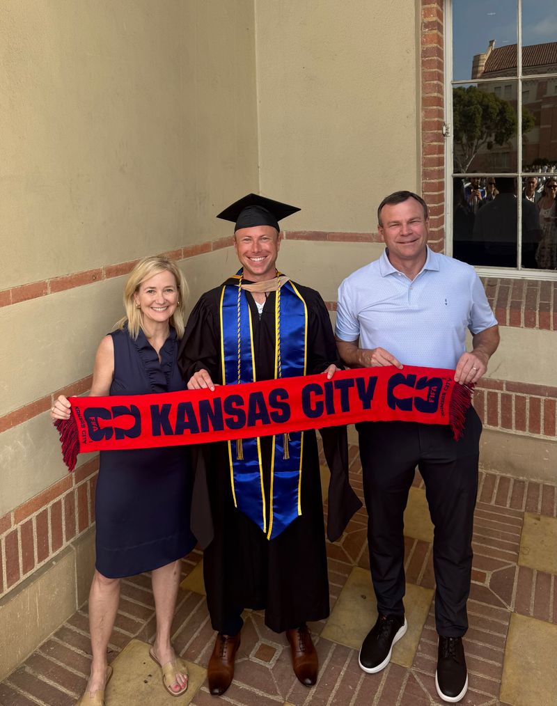A graduate in a black cap and gown with blue honors regalia holds a red banner that says Kansas City with his parents on either side of him.