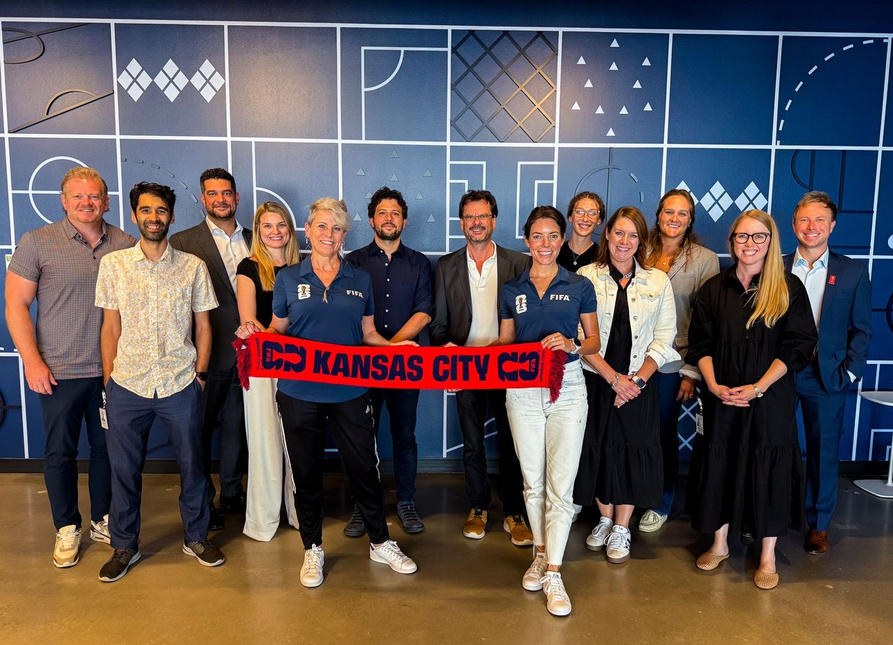 A group of 13 people stand in front of a blue wall and hold a red scarf that says Kansas City. 